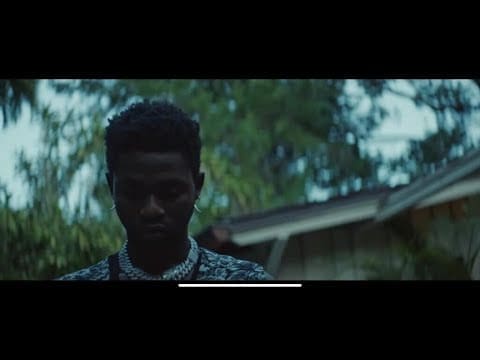 A man with short hair and a chain necklace stands outdoors in front of a house, surrounded by blurred greenery, evoking the vibe of Omah Lay's "I'm a Mess" Official Music Video.