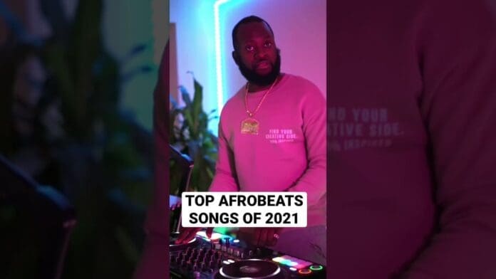 A man stands at a DJ booth with colorful lighting, wearing a pink sweatshirt and gold chain. Text on the image reads, "Top Afrobeats Songs of 2021," highlighting the best afrobeats tracks from that year.