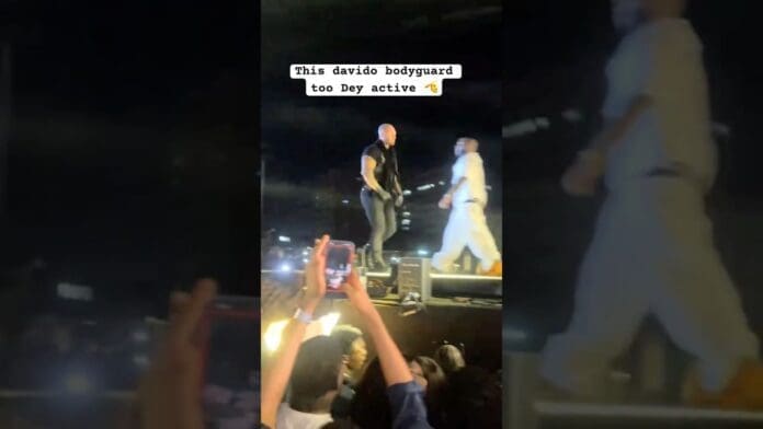 A man in black, identified as Davido bodyguard, stands alert on stage next to the afrobeats performer in white, while audience members take photos with their phones.