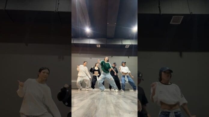 Five people in casual clothing are dancing AFRO DANCE in a studio with grey walls and a wooden floor, captured mid-move in formation as they take on an energetic dance challenge.