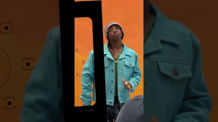 A person in a teal jacket and beanie stands against an orange background, with part of a phone screen interface visible in the foreground, capturing a vibe that echoes Afrobeats artist Bella Shmurda's "Loner" aesthetic.