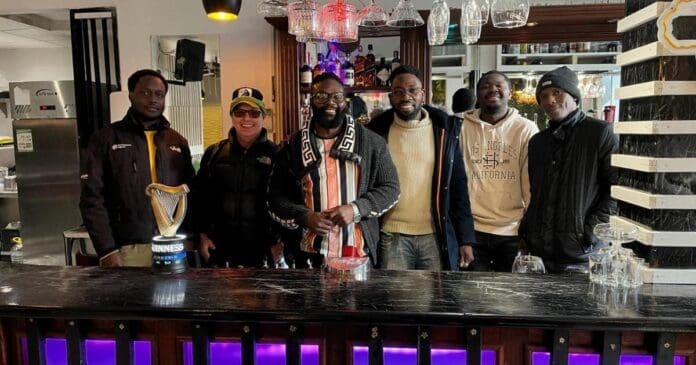 Six people stand side by side behind a bar counter with drinks and glasses, posing for a group photo in a lively Cowley Road restaurant in Oxfordshire.