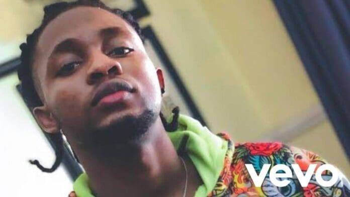 A man with braided hair and a short beard looks at the camera, wearing a colorful hoodie. The Vevo logo appears in the bottom right corner, hinting at an Official Music Video by Omah Lay.
