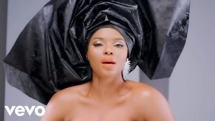 Woman wearing a large black headwrap and white earring, with bare shoulders and red lipstick, against a plain background. The "Vevo" logo in the corner hints at the iconic "Na Gode" by Yemi Alade and Selebobo.