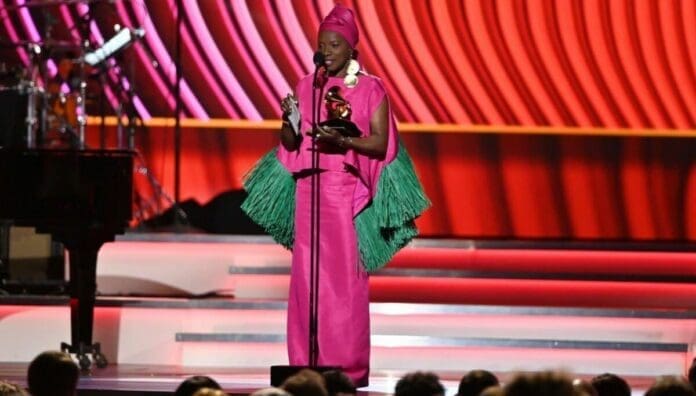 A person in a bright pink outfit with green fringe details holds a Grammy trophy while speaking on stage at an awards ceremony, marking a memorable moment in African Grammys history.
