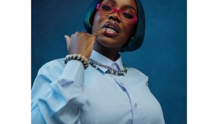 A person wearing a light blue shirt, pink glasses, and silver jewelry poses with a finger touching their lips against a blue background, channeling the vibrant style seen in OkayAfrica’s features on West African songs.