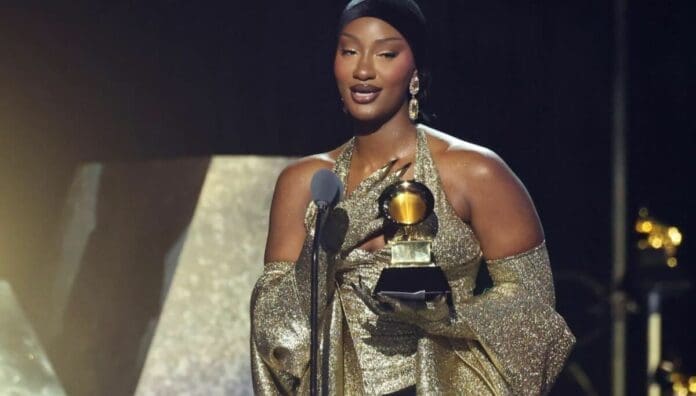 A woman in a gold dress stands on stage at the 2025 Grammys, holding a Grammy award and speaking into a microphone, celebrating the impact of African Music.