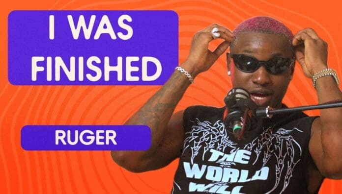 A person with pink hair and sunglasses speaks into a microphone. Text on the image reads "I WAS FINISHED" and "RUGER" on an orange background, highlighting the Afropop star’s feature on Afrobeats Intelligence.