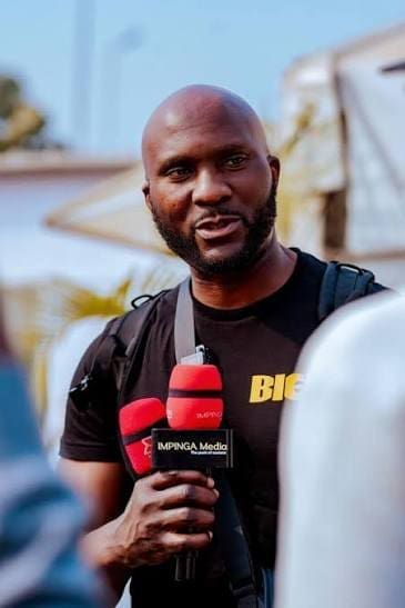 21257584772026.jpeg A man holding two red microphones and wearing a black T-shirt with "BIG" printed on it speaks outdoors during an interview about the Grammy Results.