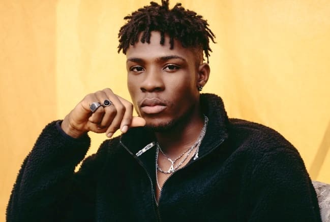 A young man with short dreadlocks and facial hair poses in front of a yellow background, wearing a black fleece jacket and multiple silver chains and rings, channeling Afrobeats style reminiscent of Joeboy.