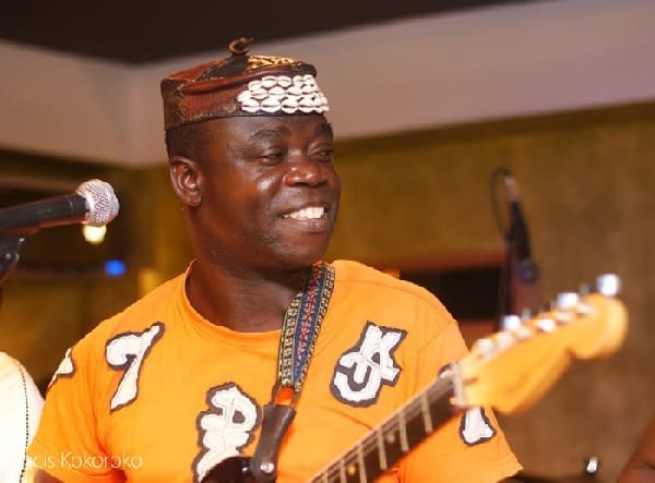 A man wearing an orange shirt and a decorated cap plays an electric guitar and smiles while performing afrobeat music on stage.