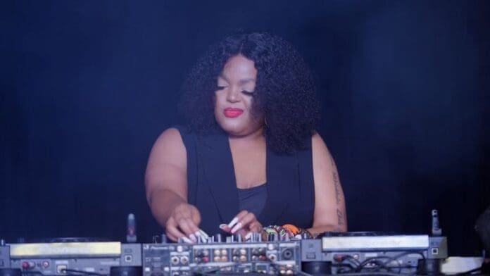A DJ with curly hair and red lipstick operates a sound mixer, standing between two DJ turntables against a dark background, showcasing the vibrant Amapiano music genre from South Africa.
