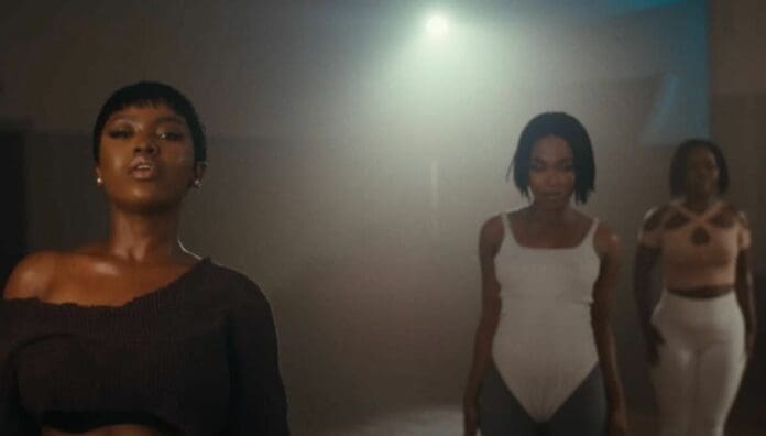 Three women in dancewear stand in a dimly lit studio, facing forward with neutral expressions, as the latest Afrobeats songs from OkayAfrica play softly in the background.