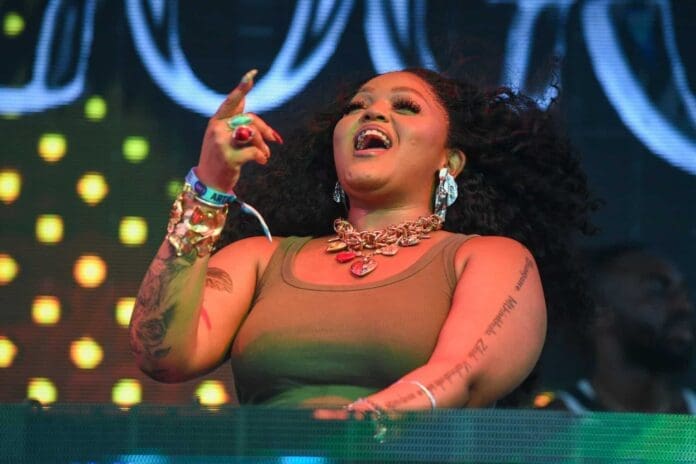 A woman with long curly hair performs on stage, wearing large earrings, bracelets, and a tank top, with colorful lights in the background—channeling DBN Gogo as she aims to break a record with an electrifying 24-hour set.