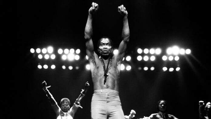 A shirtless man stands on stage with both fists raised in the air, flanked by two other performers, as bright stage lights shine behind them—a powerful tribute to Fela Kuti and his enduring music legacy.
