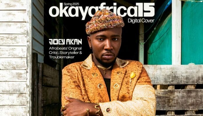 A man in a patterned jacket and knit hat stands in front of a rustic wooden wall for the cover of OkayAfrica magazine, Spring 2025 issue, with text introducing Afrobeats journalist Joey Akan.