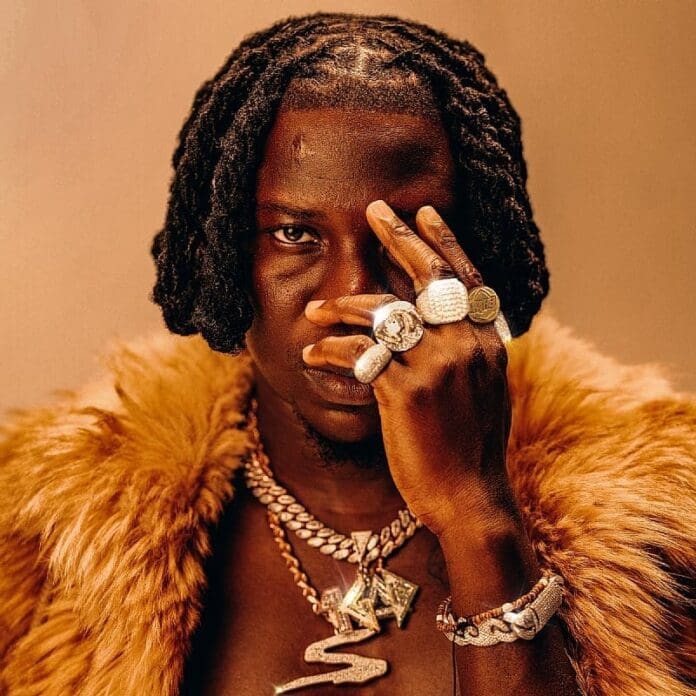 A person wearing multiple rings and necklaces, dressed in a brown fur coat, covers one eye with their hand against a neutral background—channeling Stonebwoy’s bold style as seen at the Africa Prosperity Dialogues.