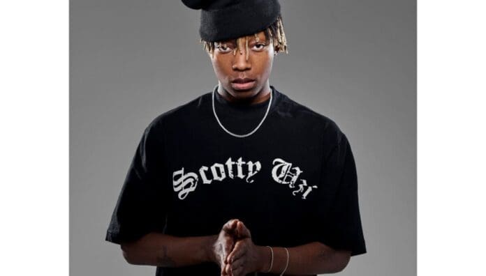 A person wearing a black beanie, black t-shirt with "Scotty Chi" text, and a silver necklace stands against a plain gray background with their hands clasped, channeling the cool vibes of South Africa’s Amapiano scene.