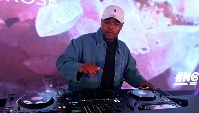 A DJ wearing a white cap and denim jacket performs Amapiano songs on a mixing console, with a colorful background display behind him at the OkayAfrica 2024 event.