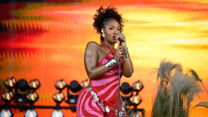 A woman in a red and pink striped outfit sings afrobeats into a microphone on stage, with stage lights behind her and tall decorative grasses to the side, echoing Nigeria’s global power in music.