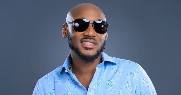 A man wearing dark sunglasses and a light blue floral shirt channels Afrobeats style as he poses against a plain grey background.