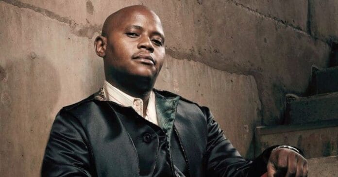 A man in a dark jacket sits on concrete stairs against a rough, beige wall, looking confidently at the camera—his style reminiscent of classic Kwaito vibes and the energy of Joe Nina’s iyano fusion.