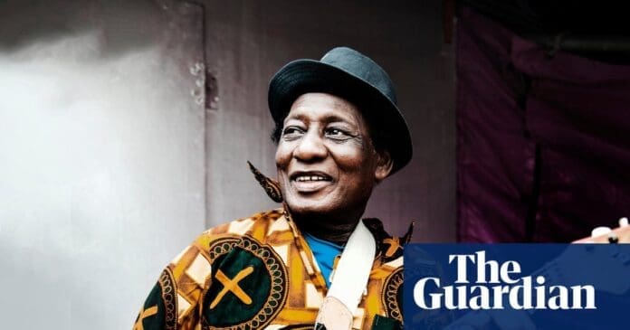 A man wearing a patterned shirt and a hat smiles while holding a guitar, channeling Ghanaian highlife vibes. The Guardian logo appears in the corner.