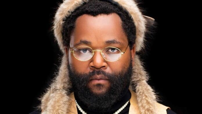 A man with a full beard wears round glasses, layered necklaces, and a fur hat with animal ears, embodying the eclectic style seen in Sjava's new album Inkanyezi Nezinkanyezi, against a black background.