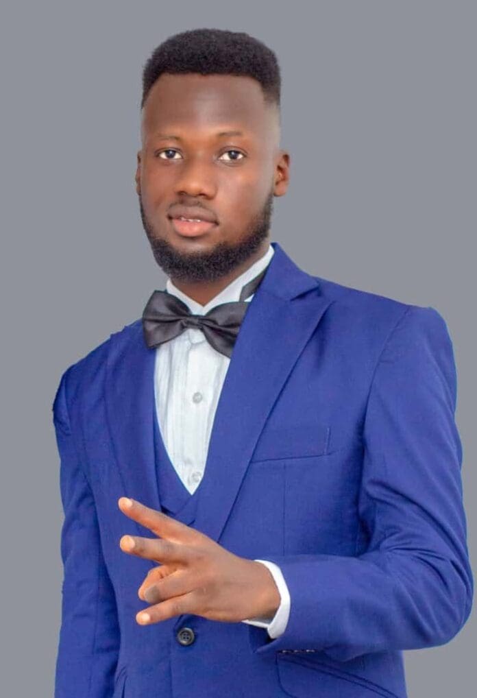 A Ghanaian artist in a blue suit and bow tie stands against a plain gray background, holding up three fingers with his right hand.