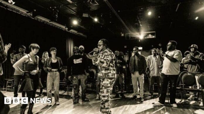 A performer with a microphone stands in the center of a room, surrounded by an audience, under stage lighting, channeling the energy of Fela Kuti and the vibrant rhythms of Afrobeat at a Hull exhibition.