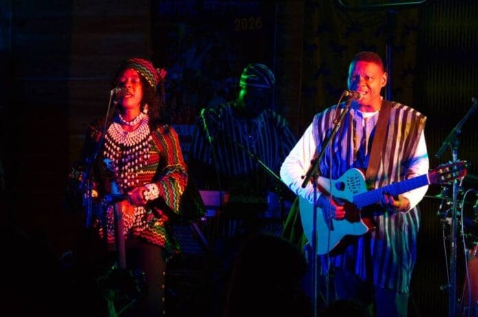 Two musicians in traditional African attire perform on stage at the Yukon African Music Festival; one plays guitar and sings while the other sings into a microphone. A keyboard player is visible, highlighting the talent of global artists.