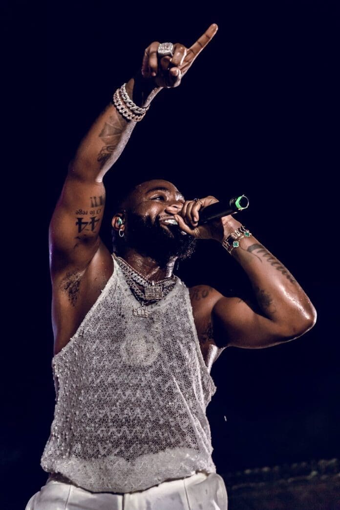 A man wearing a sleeveless mesh top and jewelry performs on stage, holding a microphone and raising one arm with his index finger pointed upwards—a striking moment among African Artists who have performed in the most countries, as featured on GhanaWeb.