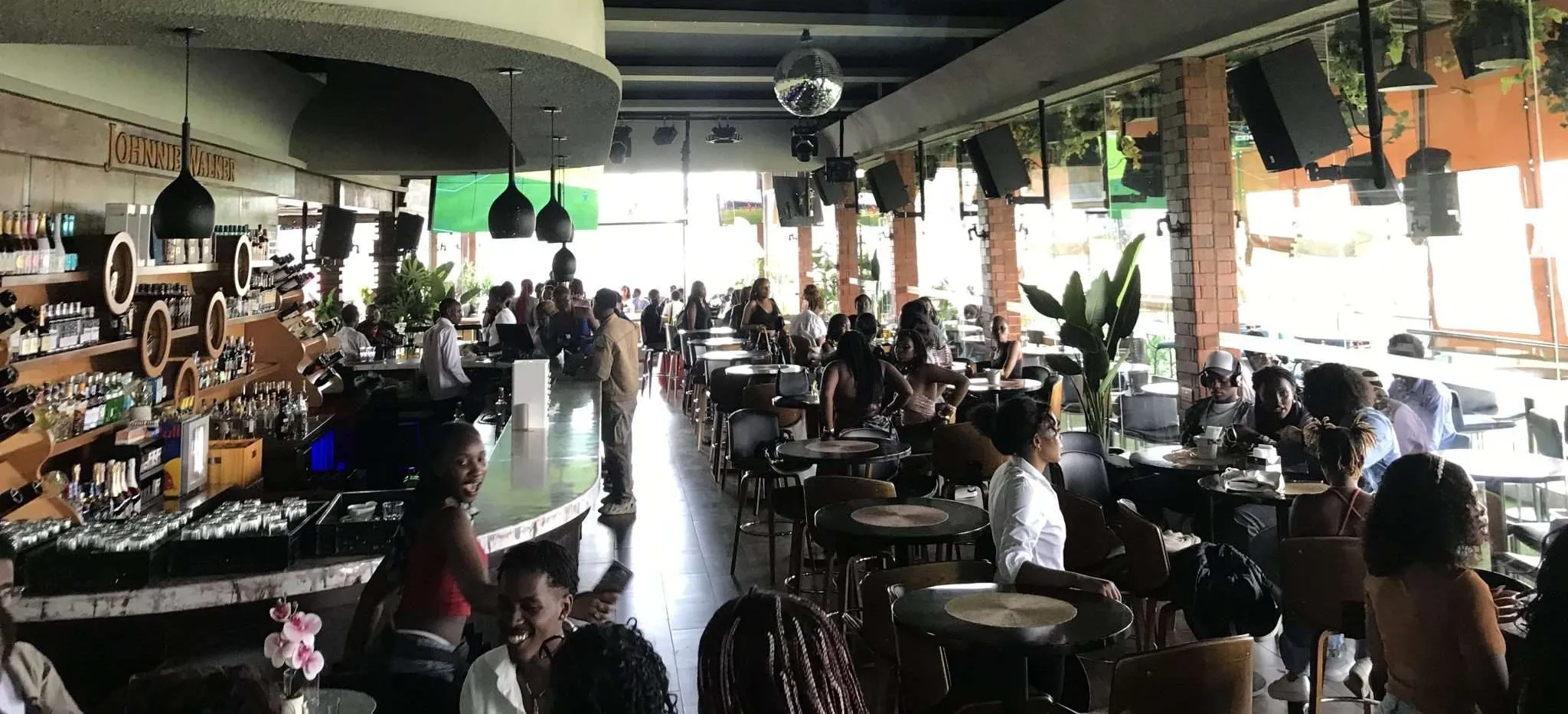 A modern restaurant and bar, Orchid Lounge, with people seated at tables and the bar, featuring hanging lights, large windows, green plants, and a lively KenyanVibe.