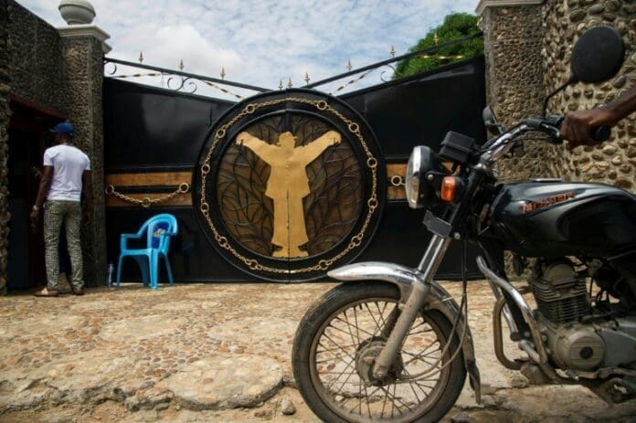 A man stands near a closed ornate gate with a gold figure design, evoking nostalgia. A blue plastic chair and a parked motorcycle rest in the foreground, while the distant echoes of Congolese rumba music linger in the air.