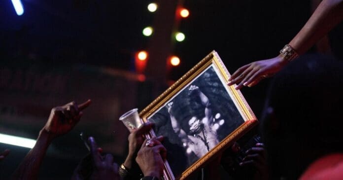 In a vibrant crowd, people hold up a framed black-and-white photograph of Fela Kuti while reaching towards a person’s outstretched hand on stage, colorful lights celebrating the legend of African music and his Grammy lifetime achievement award.