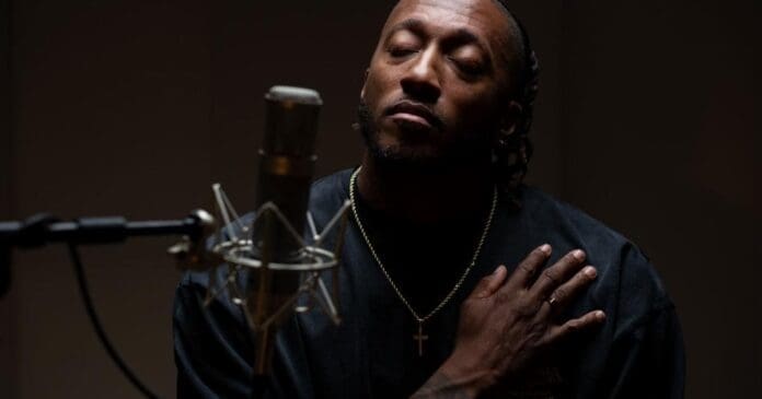 A man with closed eyes rests a hand on his chest while sitting in front of a microphone in a dimly lit setting, preparing to perform in the Christian genre.