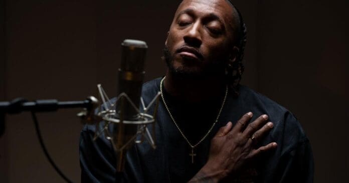 A person with eyes closed sits in front of a microphone, wearing a black shirt and a gold chain with a cross, one hand on their chest, ready to perform heartfelt Christian music.