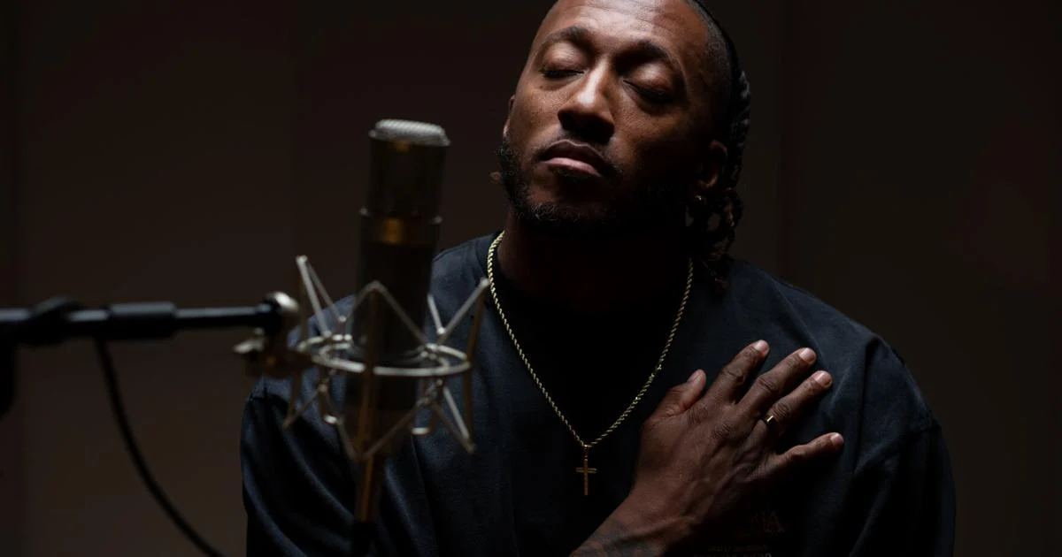 A man wearing a cross necklace sits in front of a microphone with his eyes closed and one hand resting on his chest, channeling the soulful energy of emerging artists in rap and Afrobeats.