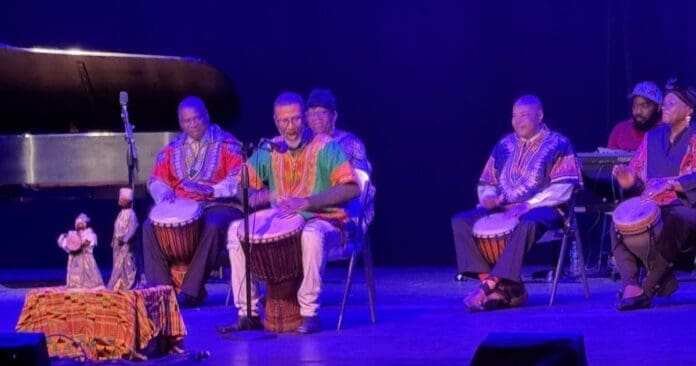 A group of musicians in colorful attire sit on stage at McGregor Hall, playing drums and filling the space with music, as a piano and a table with covered objects rest nearby under purple lighting.