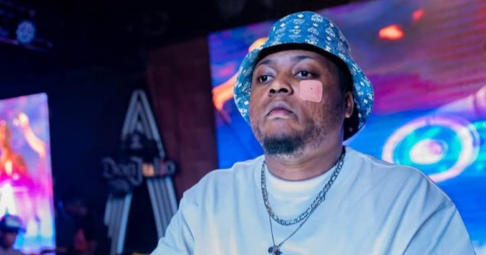 A man wearing a blue patterned bucket hat, white shirt, and silver chain with a bandage on his left cheek stands indoors with colorful lights in the background, capturing a one-man show vibe.