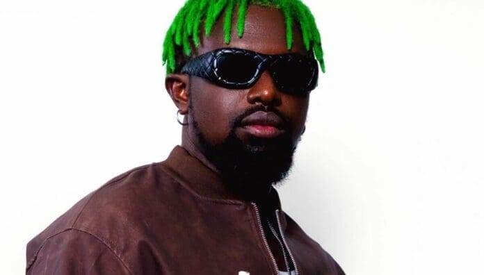 A person with bright green hair, wearing dark sunglasses and a brown jacket, poses in front of a plain white background—channeling the bold style of an Afrobeats songwriter featured on OkayAfrica.