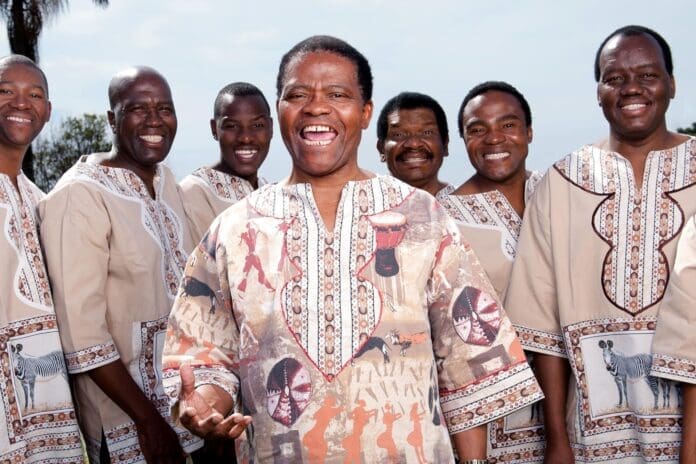 A group of seven men from Ladysmith Black Mambazo, wearing traditional patterned shirts, stand outdoors, smiling and looking toward the camera—celebrating their new album release.