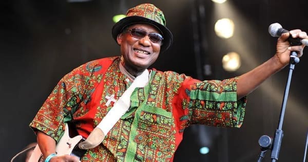 Ghanaian highlife musician Ebo Taylor smiles in a colorful patterned outfit and hat, holding a guitar and standing next to a microphone on stage.