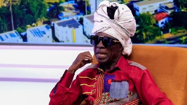 An older man wearing a white turban, dark sunglasses, and a patterned red shirt sits on an orange chair with a cityscape behind him, exuding the cool rhythms reminiscent of Kojo Antwi featured on GhanaWeb.