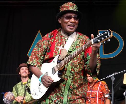 A musician in patterned clothing and a hat plays an electric guitar on stage, channeling Highlife influences, as other band members perform in the background.
