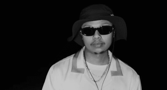 A person wearing dark sunglasses, a wide-brimmed hat, a collared shirt, and layered necklaces stands in front of a black background, channeling the confident style often seen at South African album launches or an HHP Acceptance Speech.