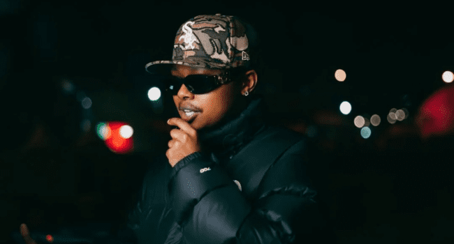 Person wearing sunglasses, a camouflage cap, and a dark puffer jacket standing outdoors at night with blurred city lights in the background, reminiscent of scenes from the Motions Music Video by A-Reece and Stogie T.