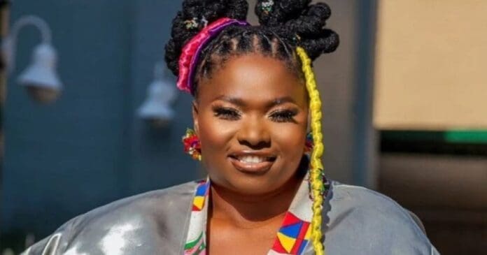 Afro-pop singer Zahara, with intricately braided hair decorated with colorful extensions, wearing a shiny silver top and patterned accessories, smiles at the camera outdoors.