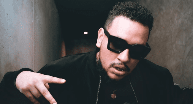 Man wearing black sunglasses and a black jacket poses with his hand raised in a dimly lit hallway, celebrating with the AKA Legacy Team and fellow AKA fans.