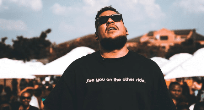 A man wearing sunglasses and a black t-shirt that says "See you on the other side." stands outdoors with people and white tents in the background, looking like he’s ready to enjoy some AKA all time hits.